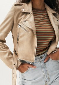 Veste de moto en daim beige avec des garnitures argentées, dotée de poches zippées, d'une coupe courte et d'une taille ceinturée. T-shirt à rayures en dessous.