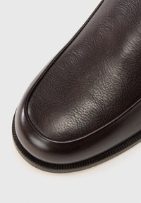 Tumepruun nahast king, millel on sile, tekstureeritud pealmine osa, ümar varvas ja õmmeldud detailid talla ääres. Lame, paindlik kummitald.