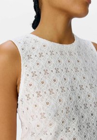 Top blanco sin mangas hecho de encaje texturizado con un patrón floral y formas de diamante, que cuenta con un escote redondo y costuras detalladas.