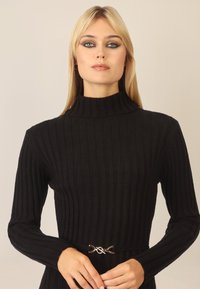Schwarzer gerippter Strickpullover mit hohem Kragen und langen Ärmeln, mit einem silbernen Hardware-Akzent an der Taille. Glatte Textur im gesamten Pullover.