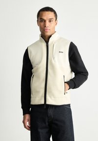 Γιλέκο από φλις σε μπεζ και μαύρο, διαθέτοντας κλείσιμο με φερμουάρ, ψηλό γιακά και λεπτομέρεια λογότυπου. Ο σχεδιασμός περιλαμβάνει μπροστινές τσέπες και αντίθετα μανίκια.