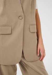 Beige tailored vest made of smooth fabric, featuring a single gold button, two front pockets, and a relaxed fit. Paired with matching pants.