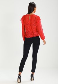 Blusa de encaje rojo con mangas acampanadas y detalle de apertura en la parte posterior, combinada con pantalones ajustados negros y tacones altos negros brillantes.