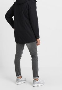 Chaqueta negra con capucha, con cierre de botones en el frente, ventilación trasera y puños ajustables, combinada con jeans grises y zapatillas blancas.