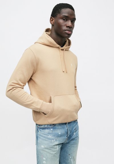 Μπεζ φούτερ με κουκούλα και τσέπη καγκουρό, με μικρό λογότυπο· φορεμένο με ανοιχτό μπλε τζιν με φθορές και λεπτομέρειες από πιτσιλιές μπογιάς.