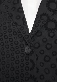 Blazer noir avec motif texturé circulaire, un seul bouton noir et un col cranté sur une chemise blanche.