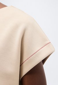 Vêtement beige à manches courtes avec un détail de couture décorative rouge sur le bord de la manche, porté par une personne à la peau foncée.