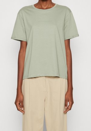 Kobieta w koszulce z krótkim rękawem w kolorze szałwi i beżowych spodniach z wysokim stanem na jednolitym białym tle.