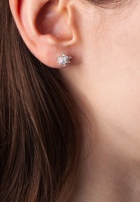 Srebrne kolczyki w kształcie płatków śniegu z sześcioma spiczastymi płatkami, z przejrzystymi kamieniami szlachetnymi, zawieszone na małżowinie usznej bez widocznych elementów mocujących lub szczegółów konstrukcyjnych.