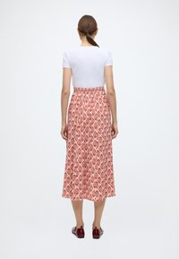 Jupe rouge à motifs avec un dessin en losanges, taille élastique et coupe longueur cheville, associée à un top blanc ajusté et des chaussures bordeaux.