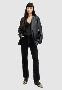 Blazer in pelle nera su un top in pizzo nero, abbinato a pantaloni in corduroy neri. Le calzature presentano un design a punta con dettagli dorati.