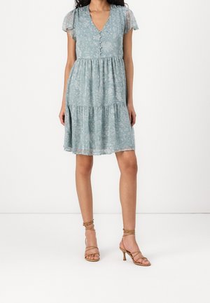 Kobieta w jasnoniebieskiej sukience w kwiaty do kolan, z krótkimi rękawami, z dekoltem w serek zapinanym na guziki oraz w beżowych sandałach na wysokim obcasie z paskami.