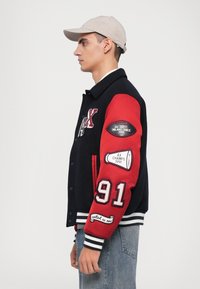 Jachetă varsity cu corp negru și mâneci roșii, având manșete ribbate albe, patch-uri brodate și numărul 91 pe mânecă.