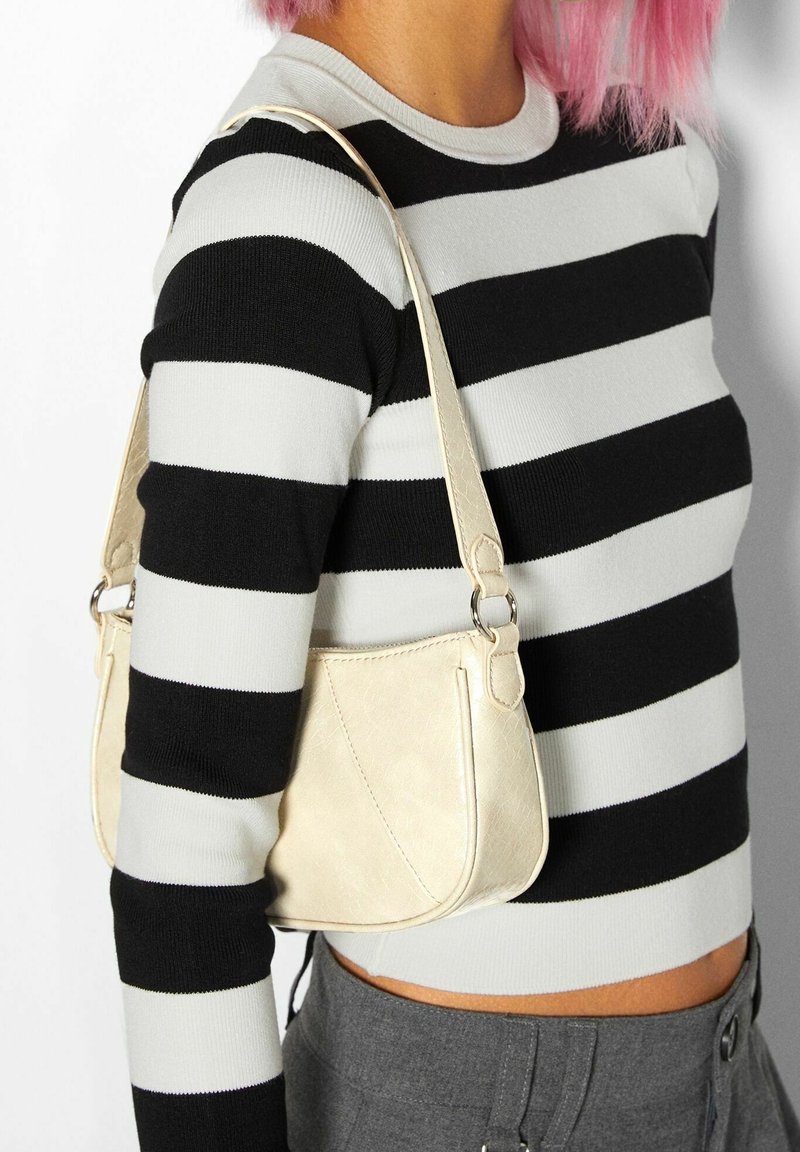 Personne aux cheveux roses portant un pull rayé noir et blanc, un pantalon gris, et un petit sac beige porté sur l'épaule droite.