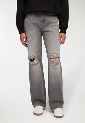 Jeans in denim grigio strappati e a vita alta con una vestibilità rilassata; presentano grandi toppe sfilacciate sulle ginocchia e un leggero sbiadimento; abbinati a sneaker bianche.