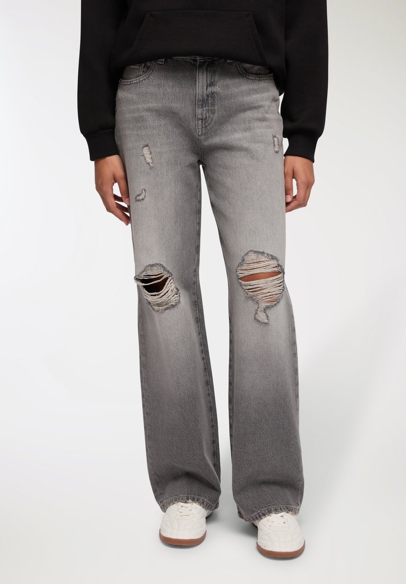 Szare, przetarte dżinsy z wysokim stanem w luźnym kroju; mają duże przetarcia na kolanach i subtelne wyblaknięcia; zestawione z białymi trampkami.