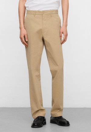 Man draagt beige rechte broek, wit ingeslagen overhemd en zwarte nette schoenen, staand tegen een effen lichte achtergrond.
