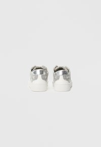 Ασημένια αθλητικά παπούτσια με λευκές σόλες, διαθέτοντας μεταλλικό προστατευτικό τακουνιού και μαλακό ύφασμα στο πάνω μέρος. Μινιμαλιστικός σχεδιασμός με χαμηλό προφίλ.