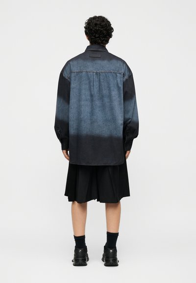 Άτομο με σγουρά σκοτεινά μαλλιά φοράει μια ξεθωριασμένη μπλε και μαύρη μακρυμάνικη μπλούζα, μαύρη πλισέ φούστα, μαύρες κάλτσες και μαύρα αθλητικά παπούτσια, στέκεται γυρισμένο με την πλάτη.