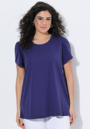 Mujer con cabello rizado y oscuro, vistiendo una blusa morada de manga corta y suelta y pantalones blancos sobre un fondo liso.