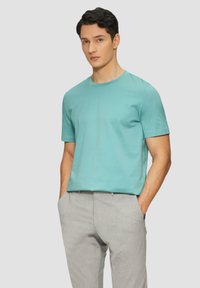 T-shirt in cotone turchese con maniche corte e scollo rotondo, abbinata a pantaloni grigi chiari con texture, dotati di tasche laterali.