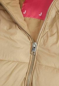 Beige quiltad jackas insida med synlig silverdragkedja och ljusrosa foder med bokstaven 'J'. Slät textur och vadderad design.