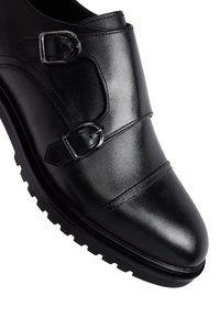 Black leather shoe featuring two buckle accents, a rounded toe, and a textured rubber sole with pronounced tread pattern.