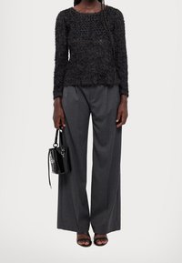Schwarzer strukturierter Pullover mit langen Ärmeln, kombiniert mit grauen weit geschnittenen Hosen. Hält eine kleine schwarze Handtasche aus Lackleder; trägt schwarze Riemchensandalen.