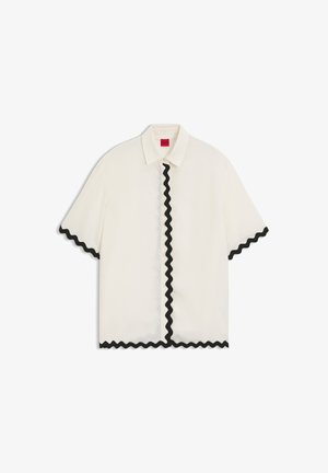 Camicia bianca a maniche corte con bordo ondulato nero lungo la linea dei bottoni, i bordi delle maniche e l'orlo, con un colletto classico e un'etichetta interna rossa.