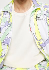 Kolorowy dres z bluzą zapinaną na zamek z zielono-fioletowo-żółtym wzorem, w połączeniu z gładkim kremowym t-shirtem pod spodem.