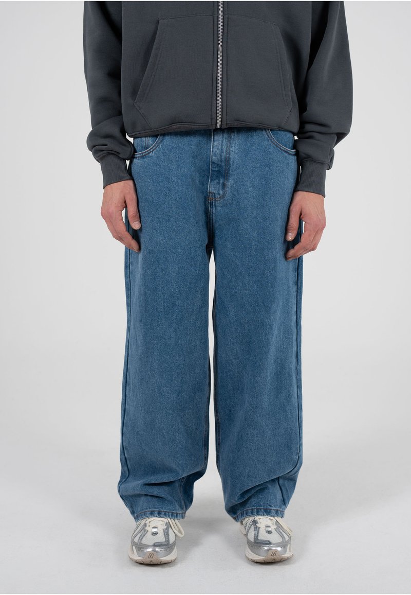 Hellblaue Jeans mit weitem Bein aus Baumwoll-Denim, mit fünf Taschen und kontrastierenden Nähten. Kombiniert mit einem dunkelgrauen Zip-Up-Hoodie und Sneakers.