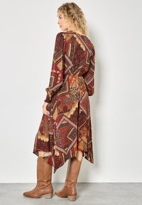 Langärmliges, mehrfarbiges Kleid mit einem geometrischen Muster in Rot, Gold und Braun. Getragen mit kniehohen, braunen Lederstiefeln.