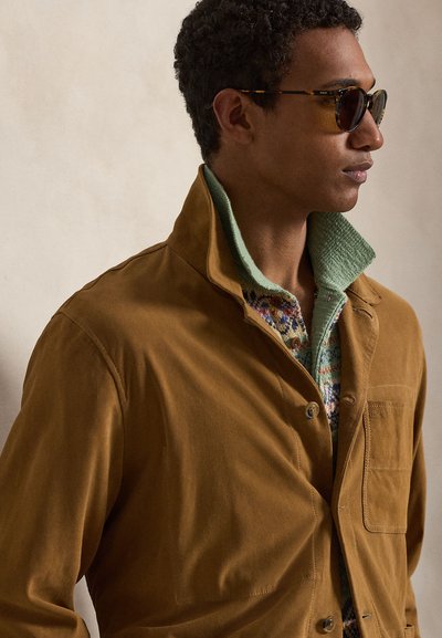 Uomo che indossa occhiali da sole tartarugati, una giacca marrone e un maglione a fantasia con il colletto verde, volto verso destra su uno sfondo semplice.