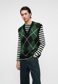 Černá a zelená vesta s kosočtverečným vzorem a výstřihem do V, nošená přes černobíle pruhované tričko s dlouhým rukávem. Pohodlný střih, texturovaná látka.