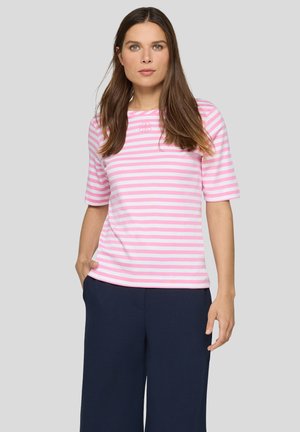 Woman wearing a pink and white striped short-sleeve shirt and dark wide-leg pants, standing with one hand in pocket against gray background.