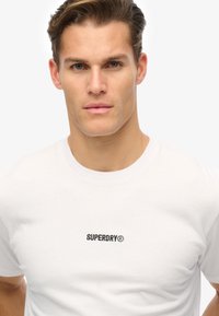 Camiseta blanca de algodón con mangas cortas y escote redondo, con un logo negro "SUPERDRY" bordado en el pecho.