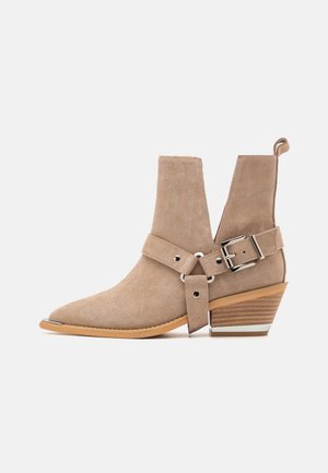Bottine en suede beige avec un bout pointu, des côtés découpés et une sangle à boucle. Présente un talon empilé avec un accent en bois et en argent.