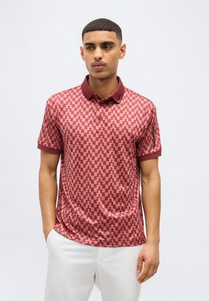 Man draagt een rood en roze poloshirt met een geometrisch patroon, een bordeauxrode kraag en witte broek, staand met handen in de zakken.