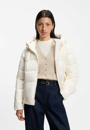 Junge Frau trägt eine cremefarbene gesteppte Jacke über einer beigen, mit Knöpfen versehenen Strickjacke und einem weißen Top, kombiniert mit dunkelblauen High-Waist-Jeans und einem schwarzen Gürtel.