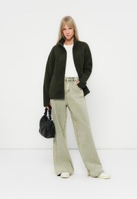 Giacca di fleece verde scuro con zip sopra una maglietta bianca, abbinata a jeans larghi verde chiaro e una piccola borsa nera. Sneakers color crema completano il look.