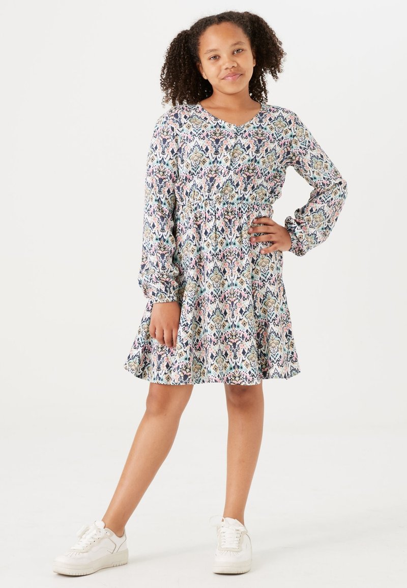 Blumendruckkleid mit langen Ärmeln, geraffter Taille und ausgestelltem Rock, in Blau-, Rosa- und Cremetönen. Kombiniert mit weißen Sneakern.