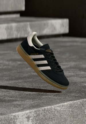 Schwarzer Sneaker aus Wildleder mit weißen Streifen und goldenem Logo, ausgestattet mit einer strukturierten Gummisohle in Kaugummifarben und einer kontrastierenden cremefarbenen Fersenschlaufe.