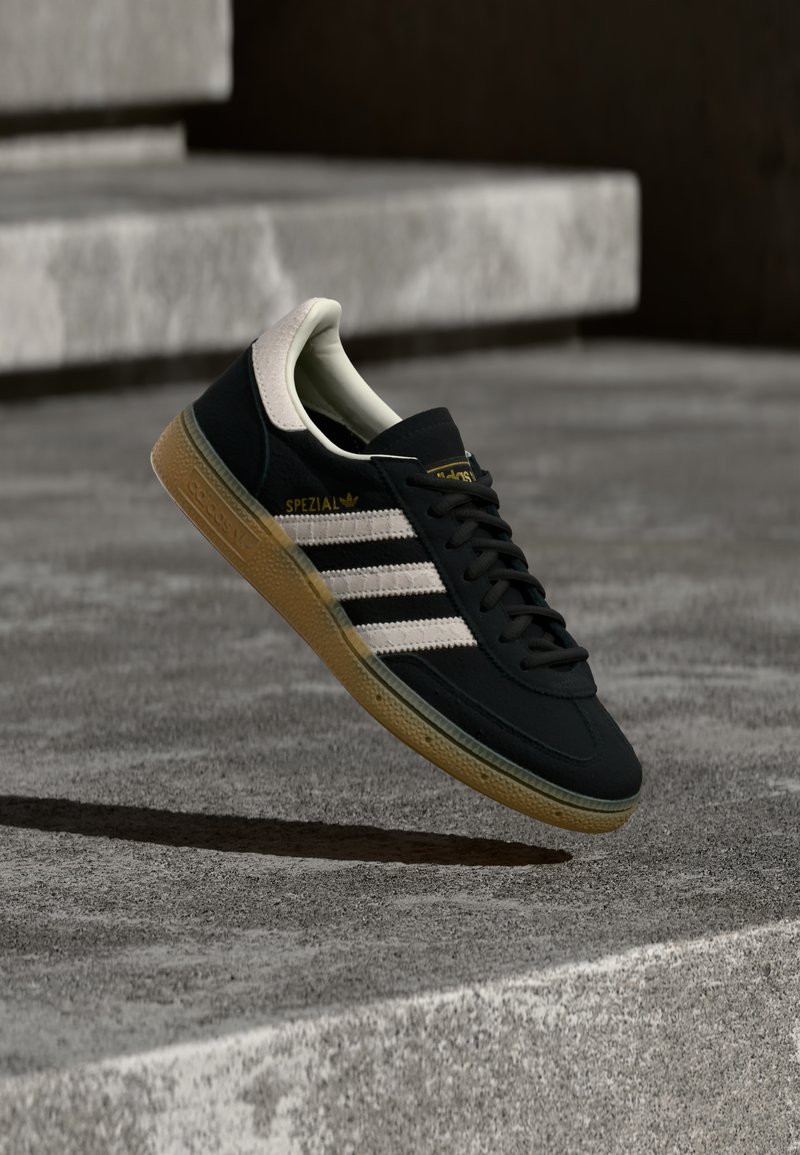 Μαύρο σουέντ αθλητικό παπούτσι με λευκές ρίγες και χρυσό λογότυπο, που διαθέτει υφασμένο πέλμα από καουτσούκ τύπου γκαμ και αντίθετη εκρού αντηρίδα στη φτέρνα.