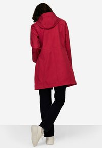 Veste imperméable rouge avec capuche, à coupe droite et détails de coutures. Portée avec un jean foncé et des baskets de couleur claire.