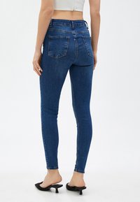 Jeans de denim ajustados en azul oscuro, con una cintura alta, dos bolsillos traseros y una textura suave. Combinados con mule de tacón negro.