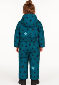 Teal winter jumpsuit met een capuchon, voorzien van zwarte en lichtblauwe sterrenpatronen. Elastische tailleband voor een goede pasvorm. Zacht, gewatteerd materiaal. Zwarte laarzen.