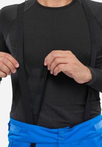 Schwarzes Langarmoberteil aus strukturiertem Stoff mit schwarzen verstellbaren Hosenträgern, kombiniert mit blauen Hosen, die einen Knopf- und Klettverschluss haben.