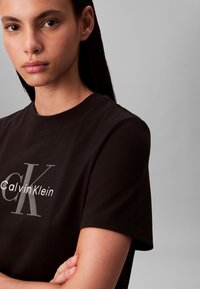 Schwarzes Baumwoll-T-Shirt mit rundem Ausschnitt, das ein weißes "Calvin Klein"-Logo und eine graue "CK"-Grafik zeigt. Kurze Ärmel, glatte Textur.