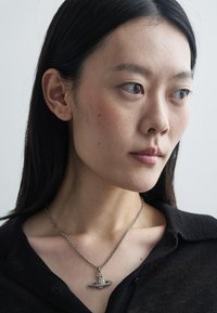 Collana d'argento con un ciondolo a forma di Saturno, caratterizzata da dettagli e motivi intricati, indossata sopra un top di tessuto nero.