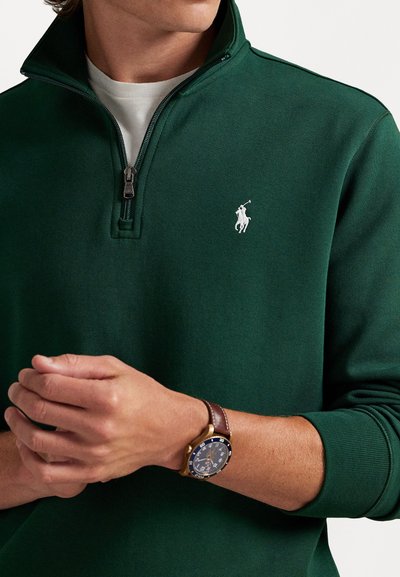 Vihreä vetoketjullinen huppari, jossa on korkea kaulus ja valkoinen polo-logo rinnassa. Yksityiskohtia ovat tyylikäs vetoketju ja teksturoitu kangas.